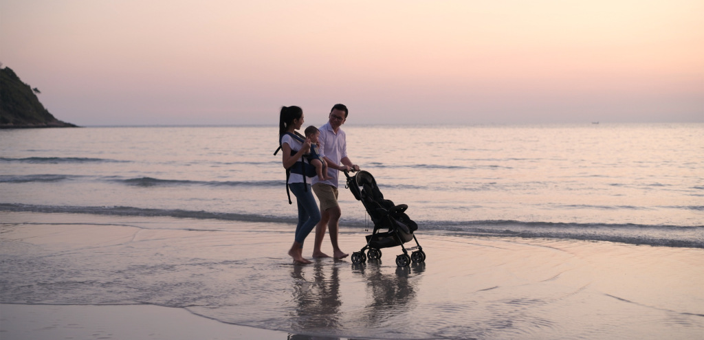 A couple walks along the beach at sunset with a stroller and baby in tow.