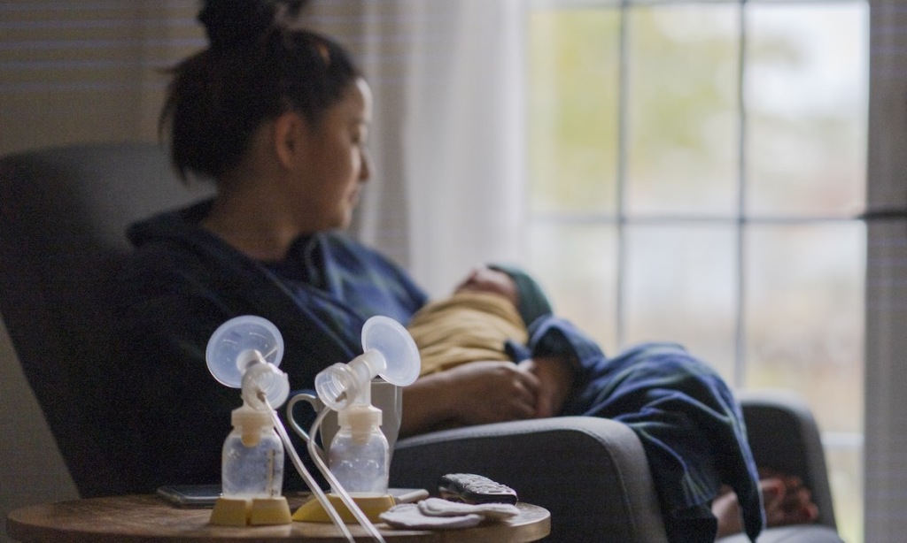 A close-up of a breast pump with a parent holding their baby in the background.