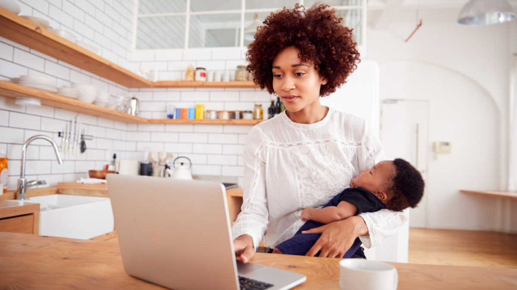 A parent holding their baby in the kitchen while on a laptop.