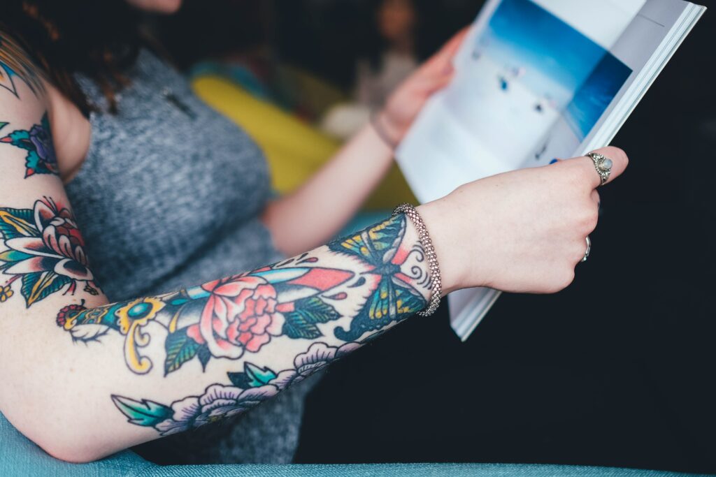 A person holds a book open to read while clothed in a sleeveless shirt with colorful, flowered tattoos on their forearms.