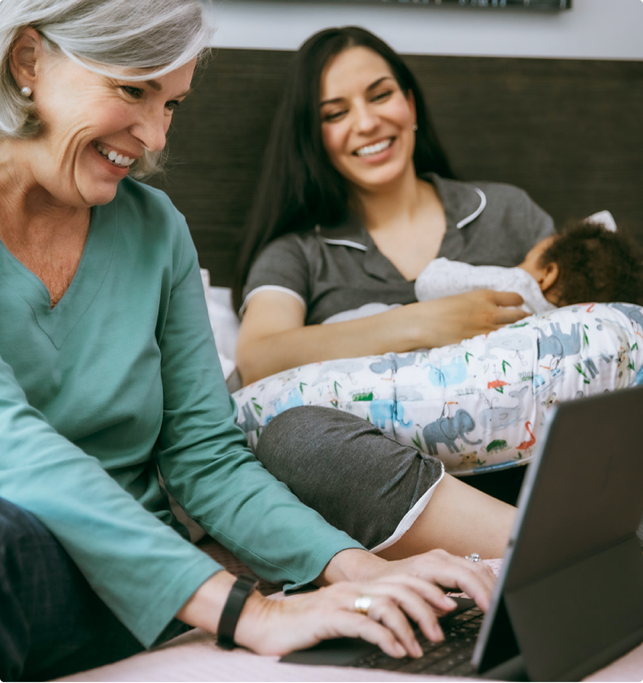 A parent breastfeeding their baby while smiling alongside another person using a laptop.