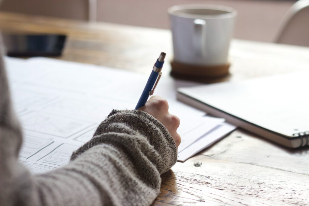 A hand slightly covered with a sweater sleeve is seen writing with a blue pen on a paper which sits on a desk.