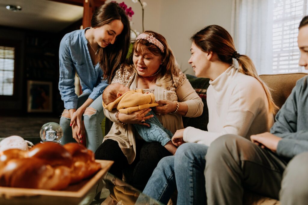 A family gathers and smiles as they look down upon a baby held in their midst by a matriarchal figure.