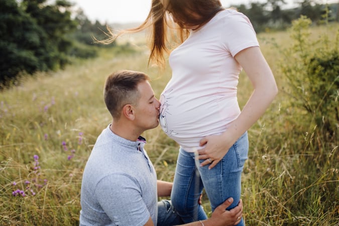 A partner kisses a pregnant parent's belly in a sunlit field, celebrating their journey into parenthood.