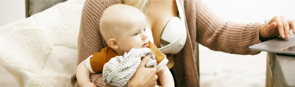 A parent holding their baby while wearing a breast pump and using a laptop.