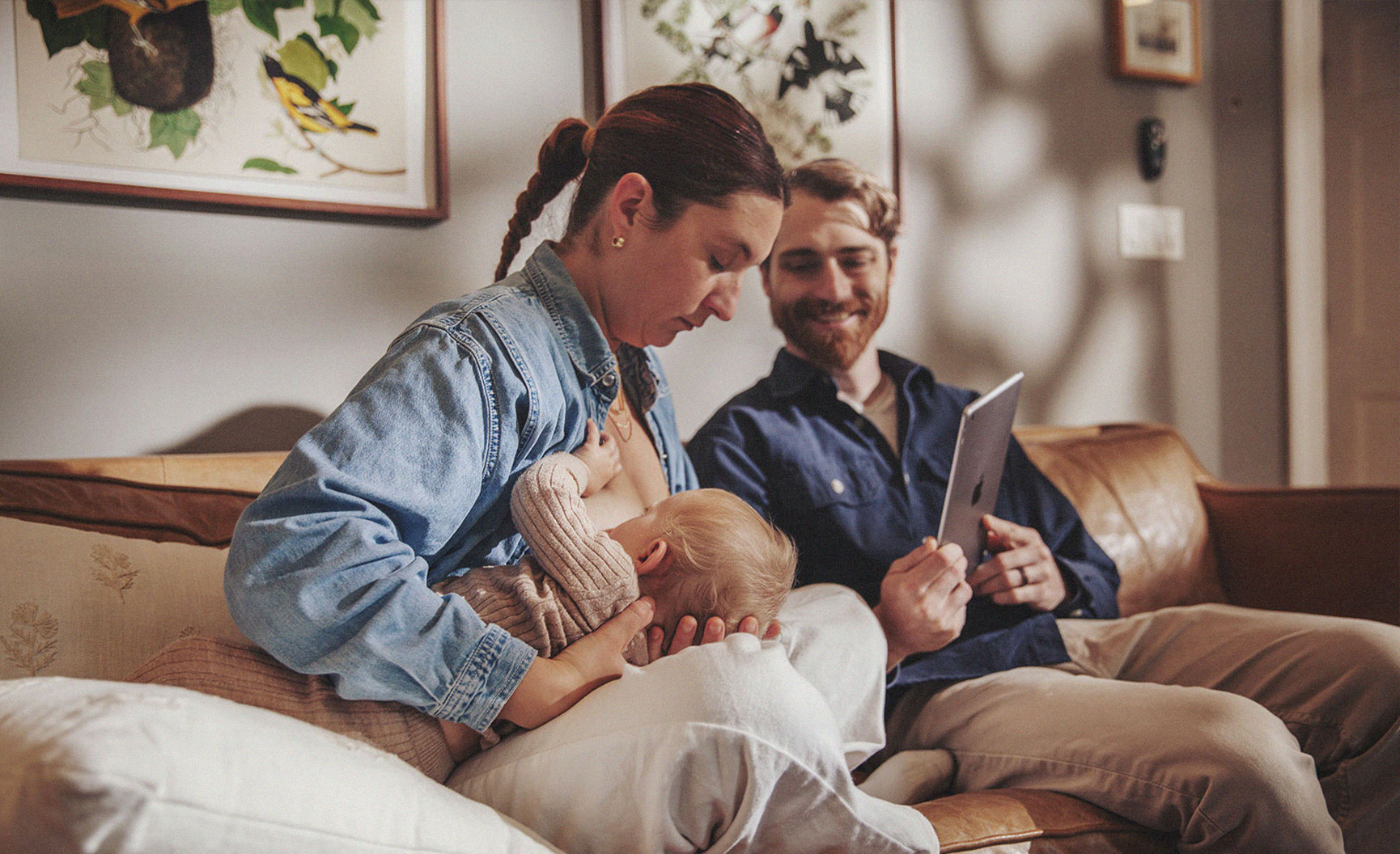A parent breastfeeding a baby while sitting on a couch, with a partner holding a tablet.