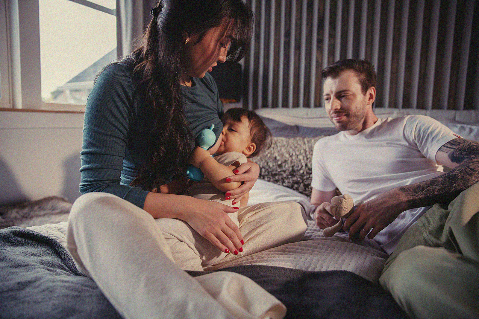 A parent breastfeeding their baby in a cozy bedroom while a partner smiles at them showing support.