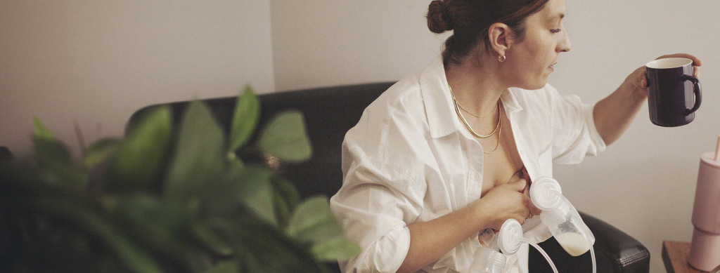 A woman using a breast pump while sitting on a couch, holding a cup of coffee in one hand.
