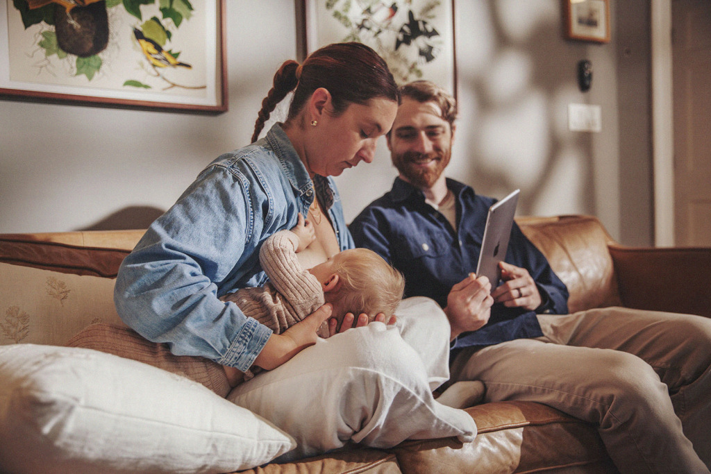 A parent breastfeeding a baby while sitting on a couch, with a partner holding a tablet.