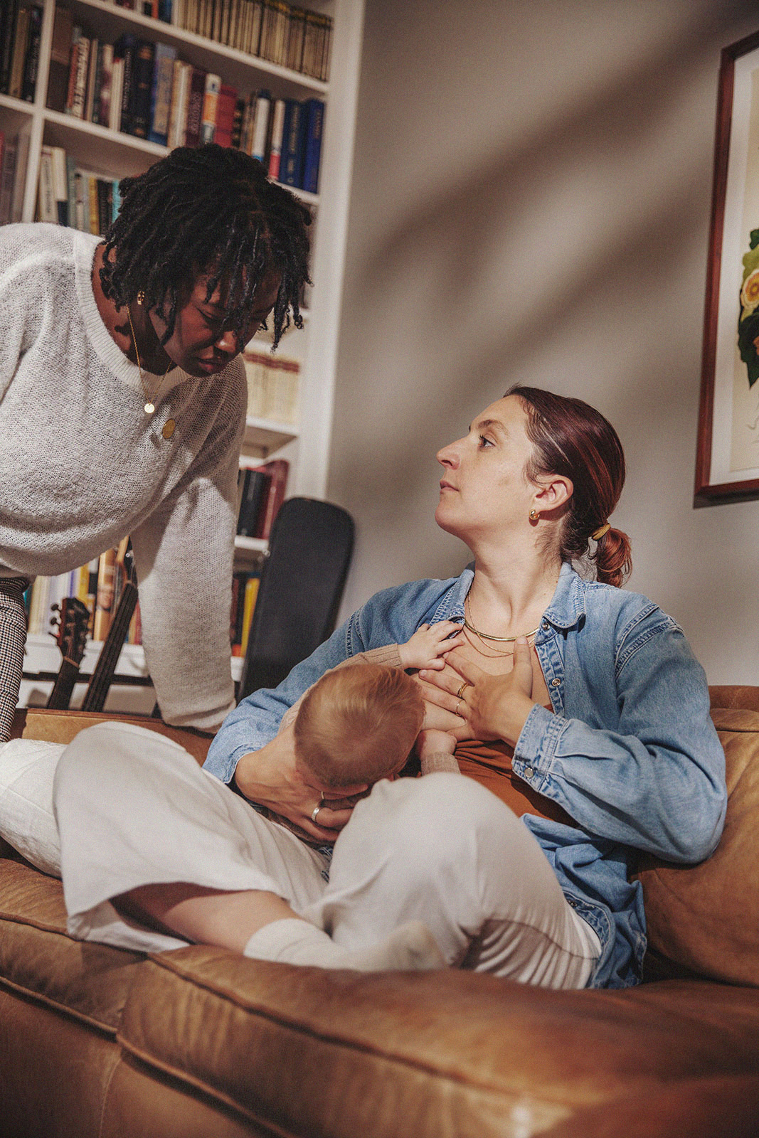 Parent breastfeeding a baby on a couch while talking to another person, showcasing a supportive moment.