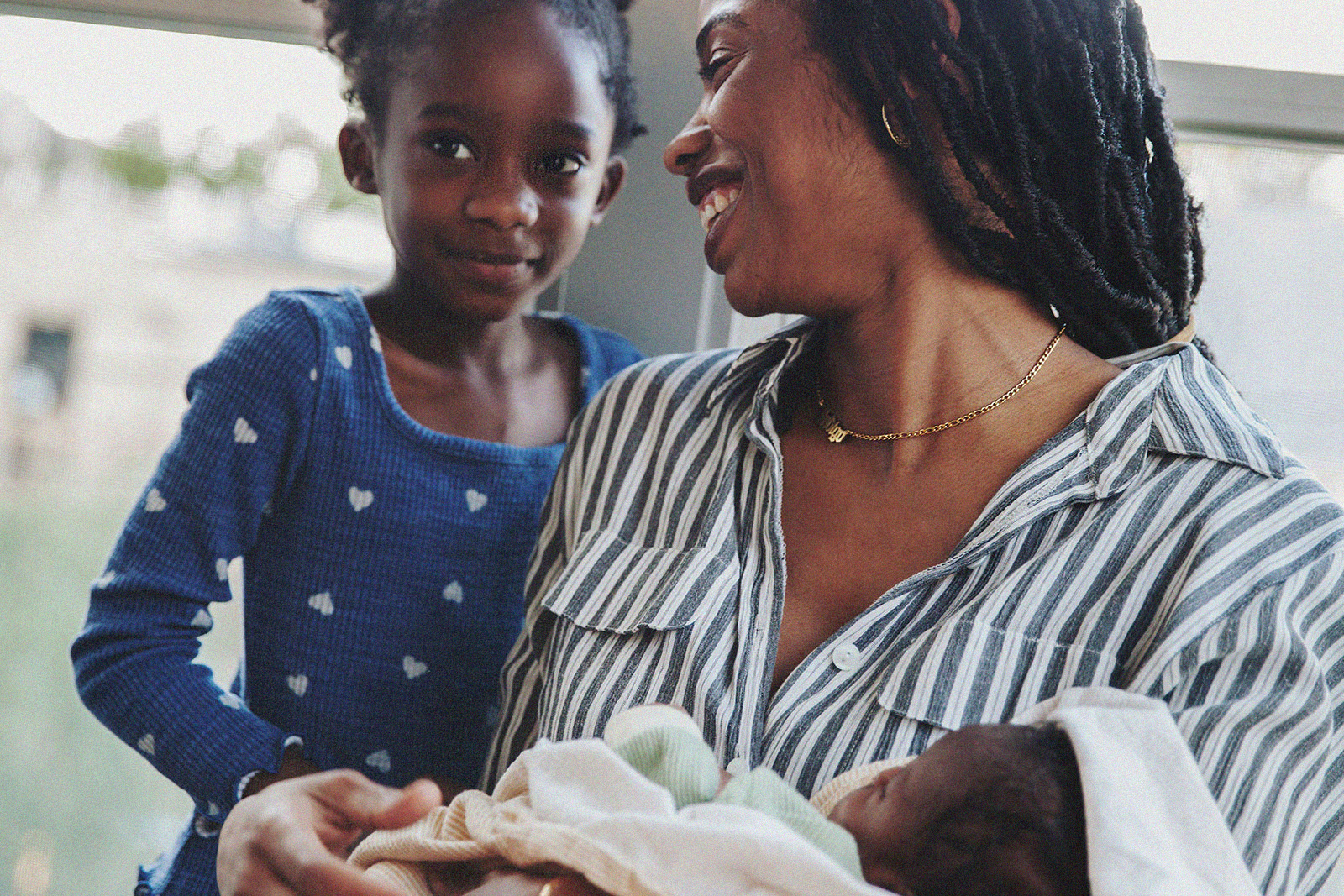 A parent bonding with her newborn while an older sibling stands by with a smile.