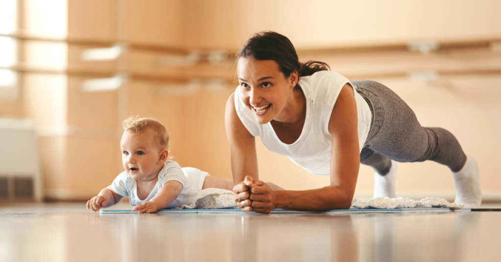 A woman holds a plank pose while smiling on a yoga mat next to a small infant.