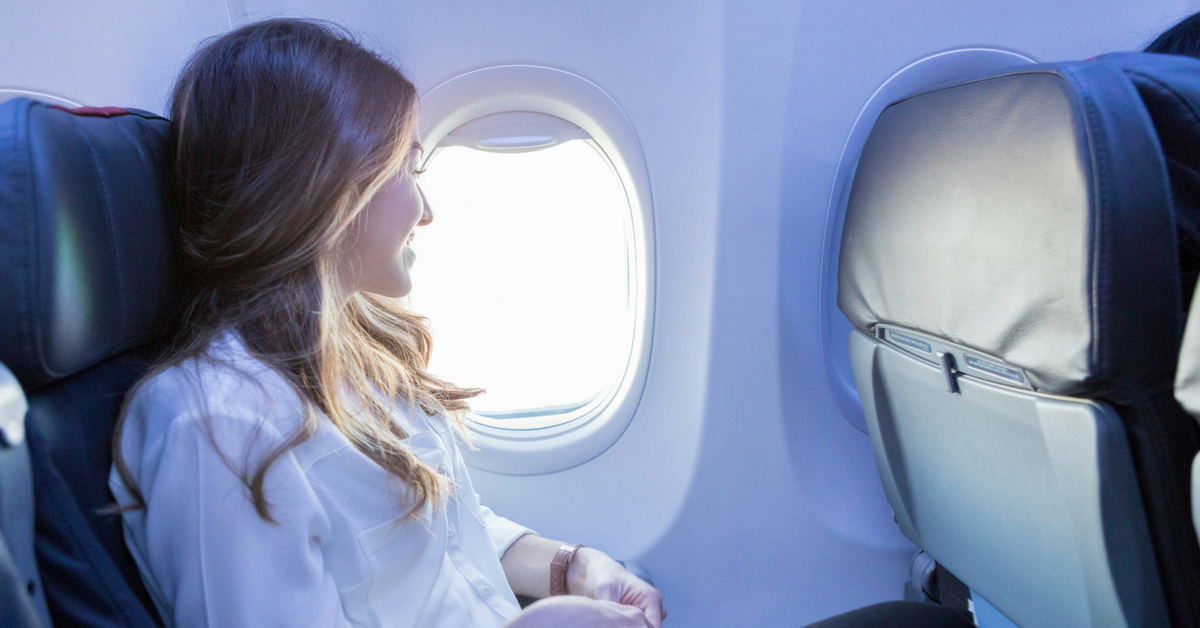 A person with long, blonde curly hair smiles as they look out of an airplane window.