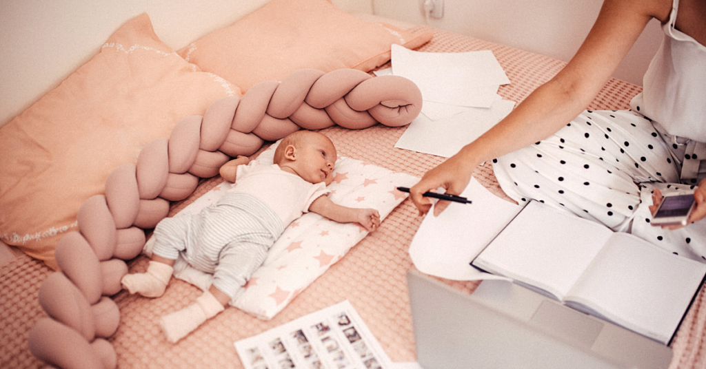 A parent holds a mobile phone while seated on the floor with paper surrounding them while their baby lays on their back on a blanket nearby
