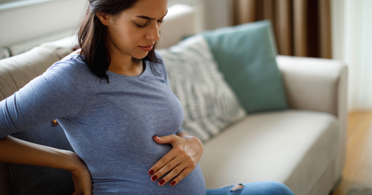 A pregnant person in a blue t-shirt sits on a sofa while touching her upper pregnant belly with her left hand.
