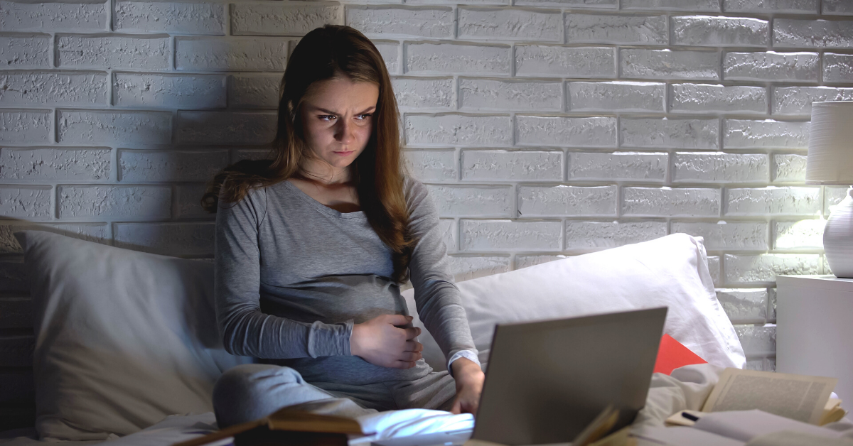 A woman with long hair worriedly looks at her laptop while sitting up in bed in the dark.