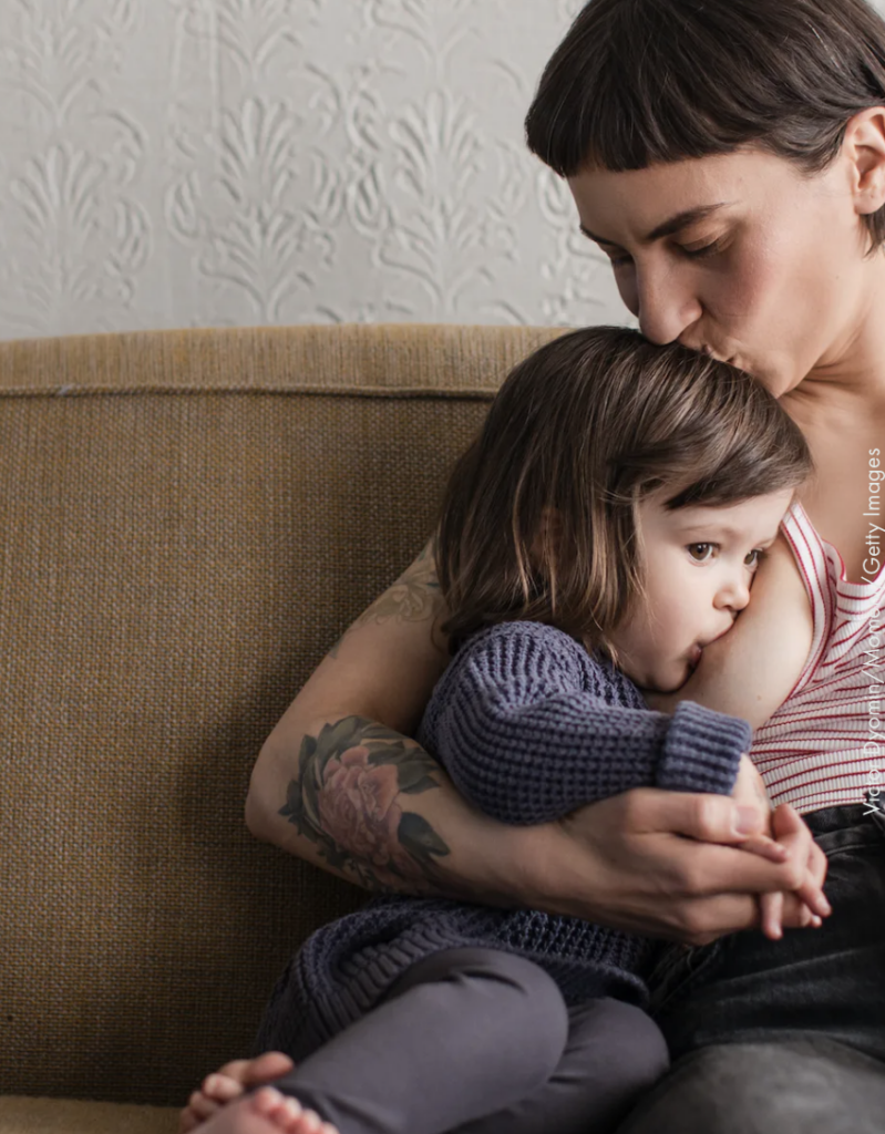 A parent breastfeeding their toddler on the couch and giving an affectionate kiss.