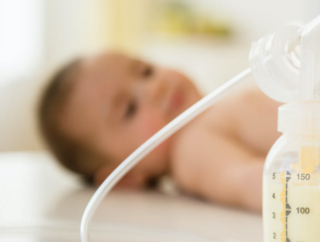 A breast pump filled with milk with a blurred infant lying in the background.