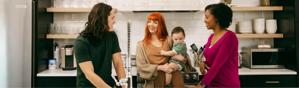A group of friends chatting in a kitchen while one holds a baby.