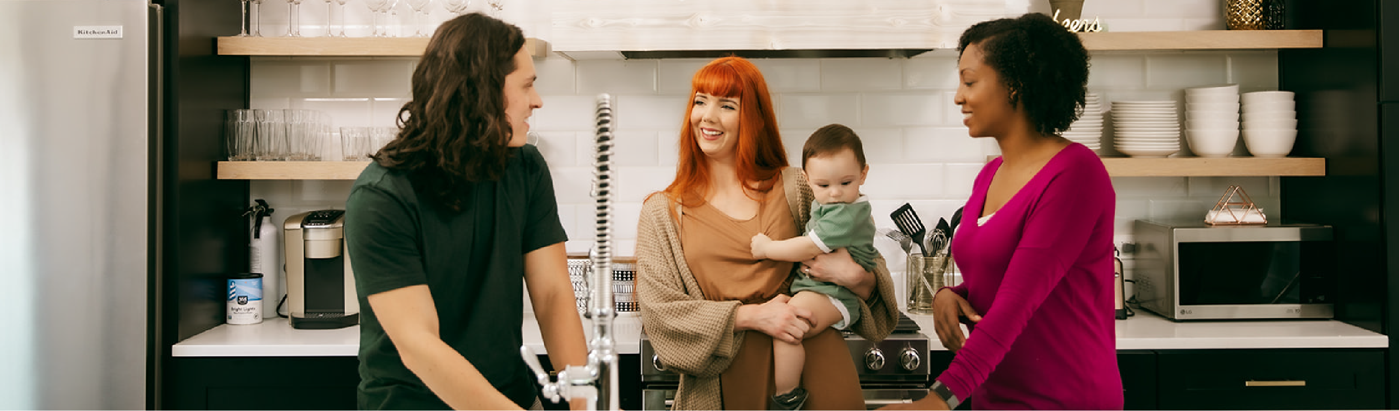 A group of friends chatting in a kitchen while one holds a baby.